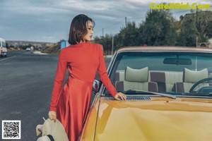 A woman in a red dress and straw hat leaning against a yellow car.