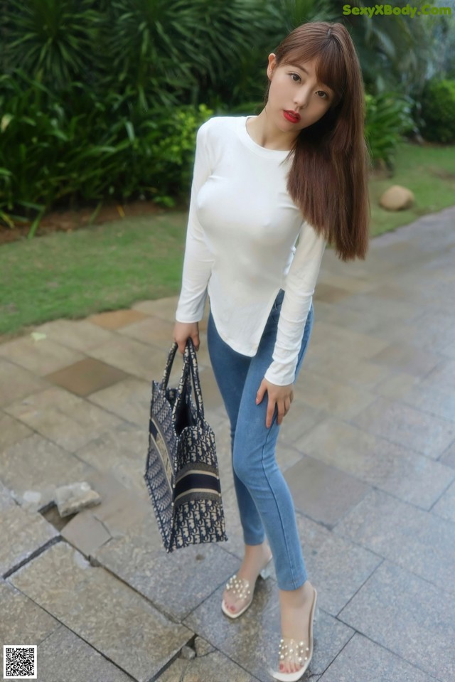 A woman in a white shirt and blue jeans holding a black bag.