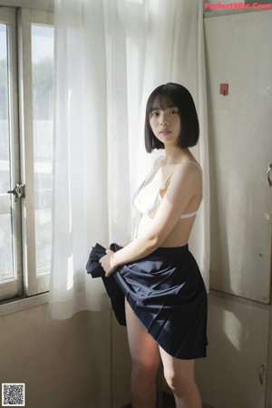 A woman in a white bra top and black skirt posing by a window.